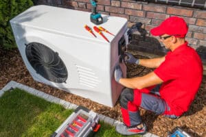 Professional Middle Aged HVAC Technician in Red Uniform Repairing Modern Heat Pump Unit.